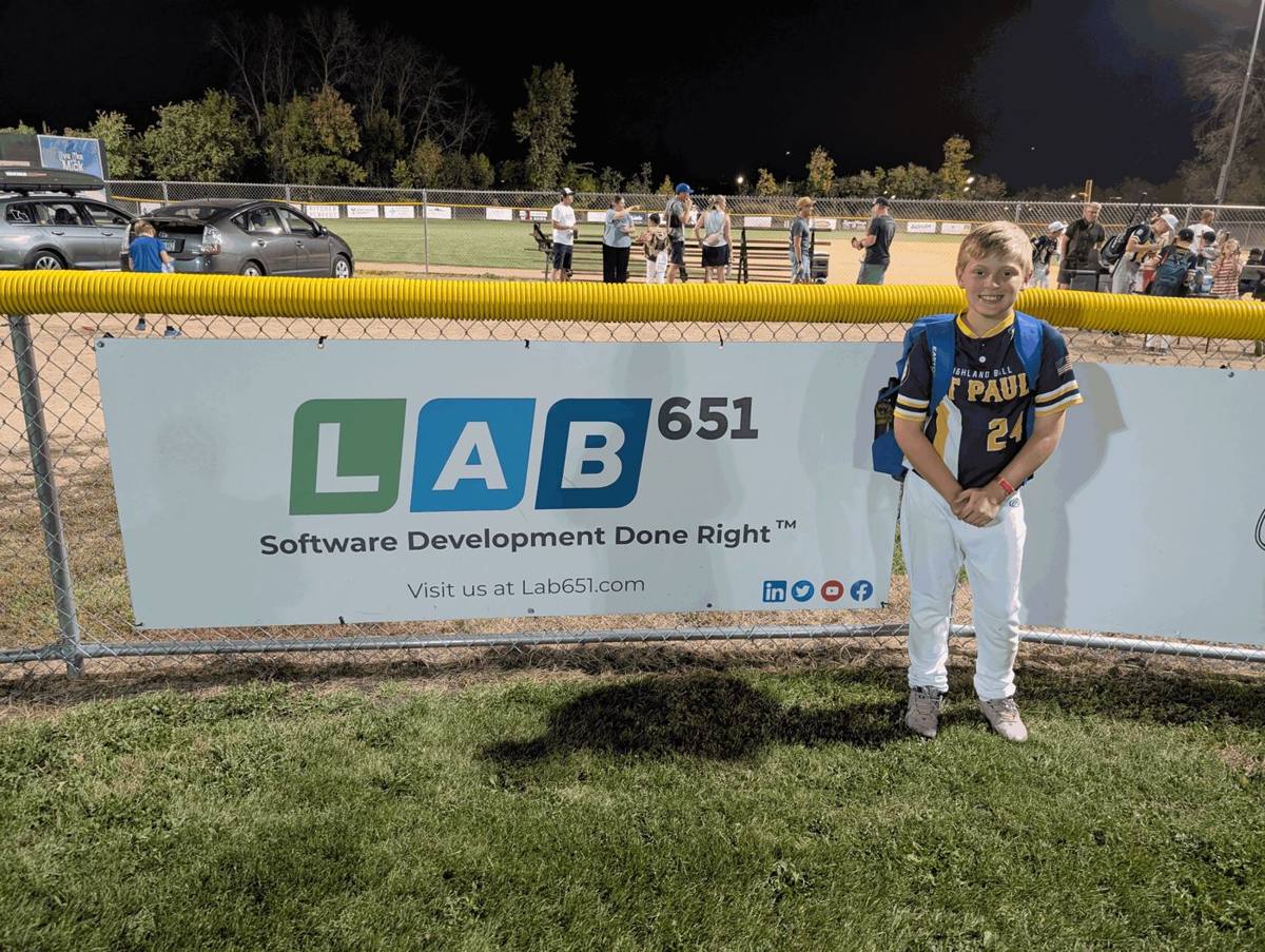 Young Highland Ball player standing next to the Lab651 sponsor banner at the ballfield